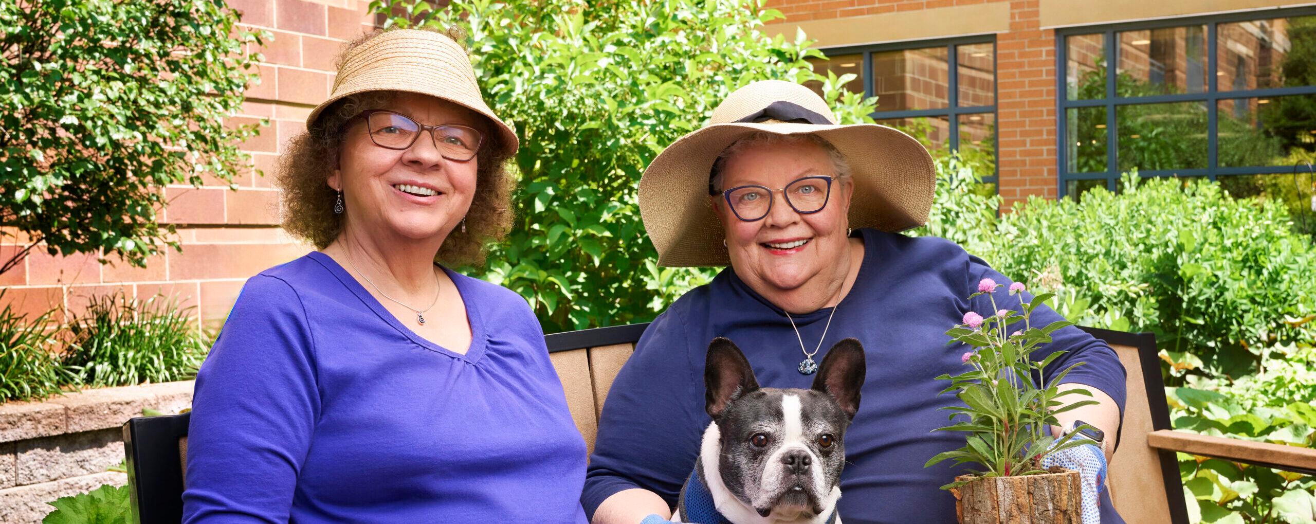 A couple sits on a bench with gardening supplies and a dog.